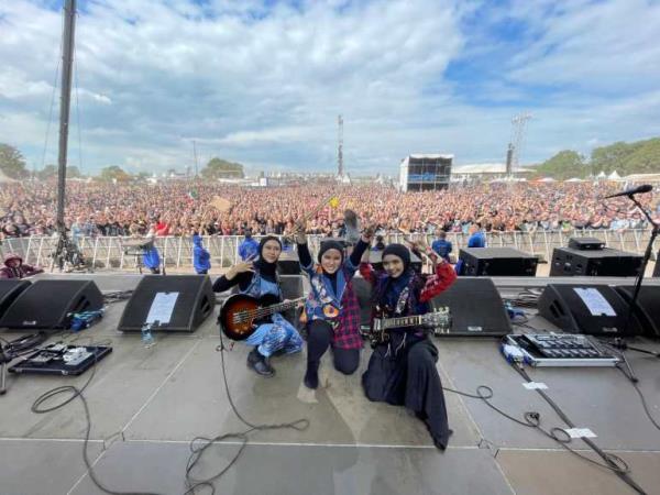 The trio on stage at Wacken Open Air, a festival in Germany/ They are posing on stage. There is a huge crowd behind them.